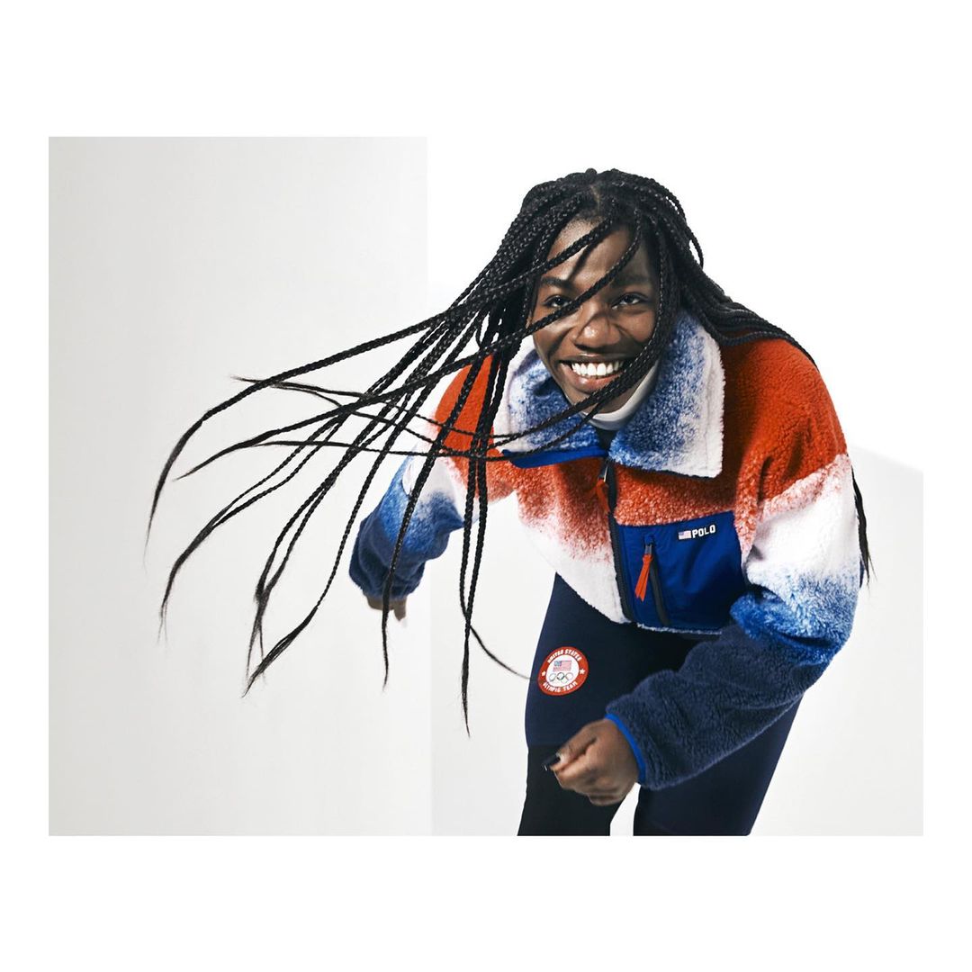 Smiling woman in red, white, and blue jacket, black leggings, poses in a photo studio with white walls, wearing braids.