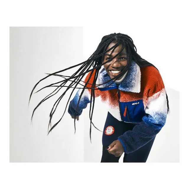 Smiling woman in red, white, and blue jacket, black leggings, poses in a photo studio with white walls, wearing braids.