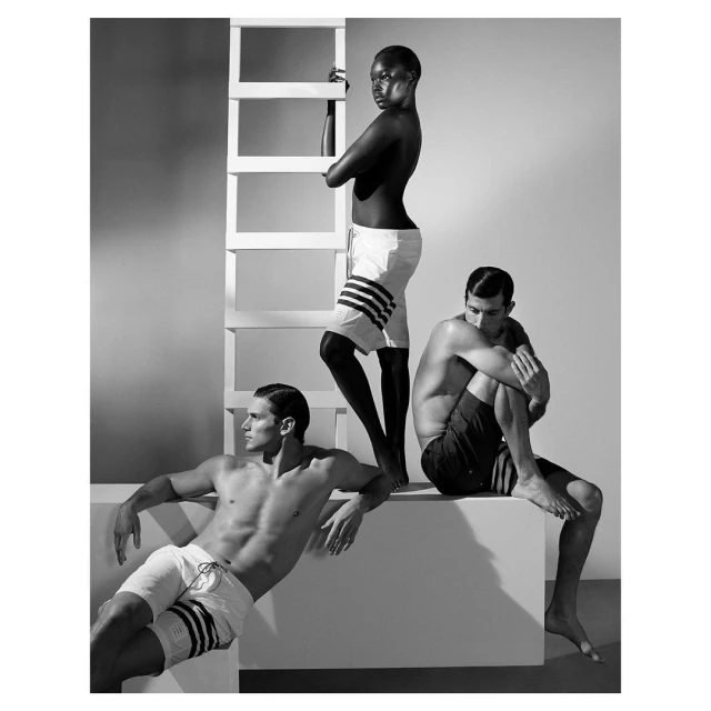 Three shirtless male fashion models in swimwear pose in a studio with a white ladder and simple set, showcasing athletic builds and swimwear.