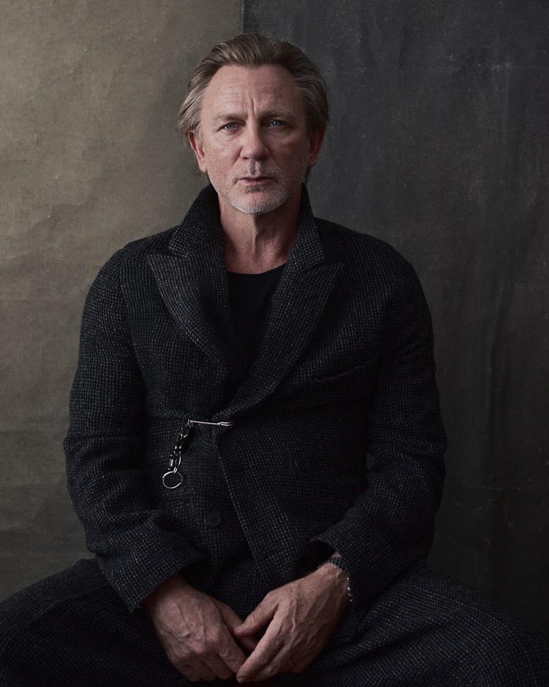 Portrait of Daniel Craig wearing a grey tweed overcoat and black shirt, seated against a textured grey and brown backdrop.