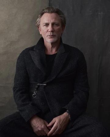Portrait of Daniel Craig wearing a grey tweed overcoat and black shirt, seated against a textured grey and brown backdrop.
