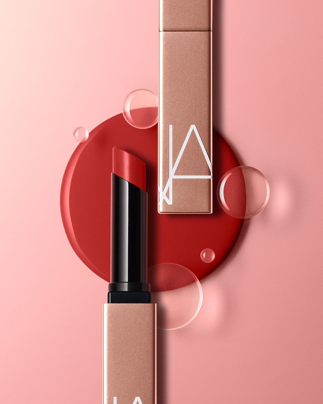 Close-up of two red lipsticks, one open and one closed, on a pink backdrop with a red circular base and transparent water droplets.