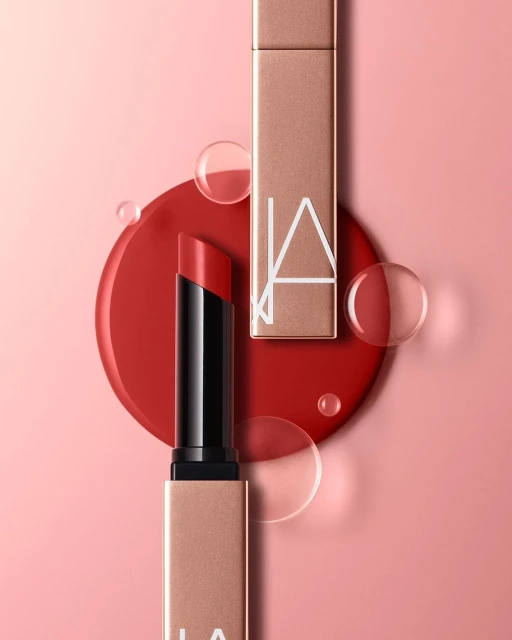 Close-up of two red lipsticks, one open and one closed, on a pink backdrop with a red circular base and transparent water droplets.
