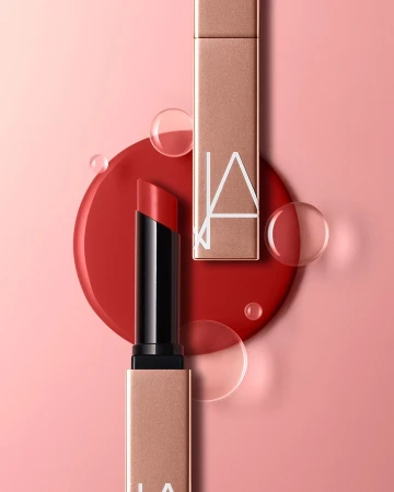 Close-up of two red lipsticks, one open and one closed, on a pink backdrop with a red circular base and transparent water droplets.