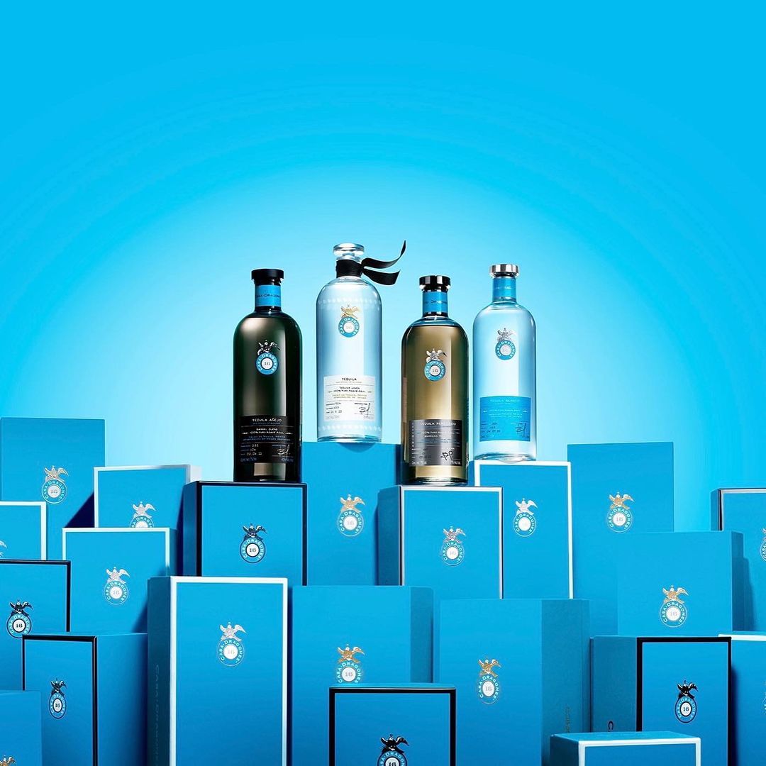Four liquor bottles in varying colors are displayed atop matching blue boxes, set against a gradient blue backdrop in a studio.