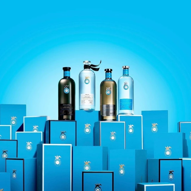 Four liquor bottles in varying colors are displayed atop matching blue boxes, set against a gradient blue backdrop in a studio.