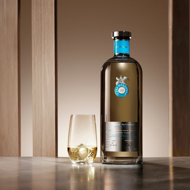 Bottle of Casa Dragones Reposado Tequila and glass with ice cubes sit on a marble surface in a studio setting with soft lighting.