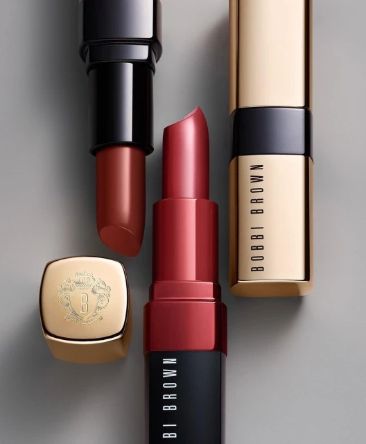 Three Bobbi Brown lipsticks in red, brown, and gold packaging are featured in a product shot against a neutral background.