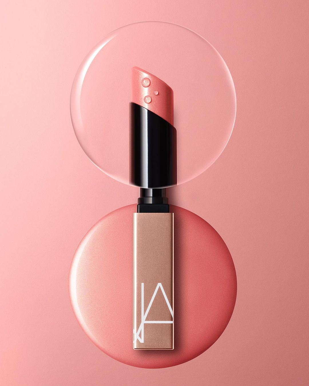 Nars lipstick product shot on coral backdrop. Beauty still life shows lipstick tube with water droplets on coral-colored lipstick.