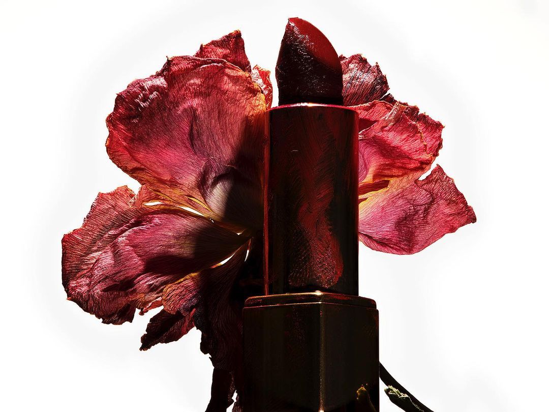Macro studio shot of red lipstick tube positioned near a wilted flower with curled, textured petals against a bright white backdrop.