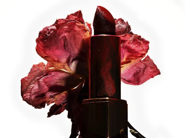 Macro studio shot of red lipstick tube positioned near a wilted flower with curled, textured petals against a bright white backdrop.