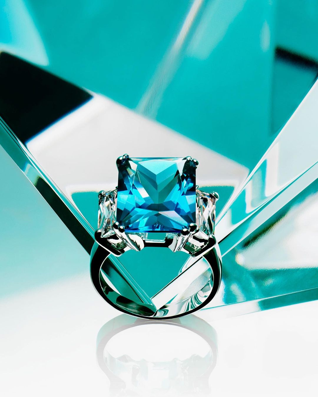 Elegant silver ring featuring a large, square-cut blue gemstone, flanked by smaller clear stones on a glossy white surface.