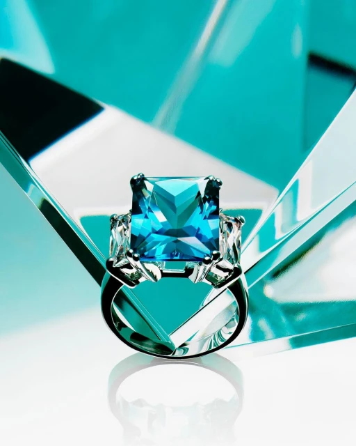 Elegant silver ring featuring a large, square-cut blue gemstone, flanked by smaller clear stones on a glossy white surface.