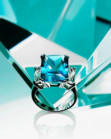 Elegant silver ring featuring a large, square-cut blue gemstone, flanked by smaller clear stones on a glossy white surface.