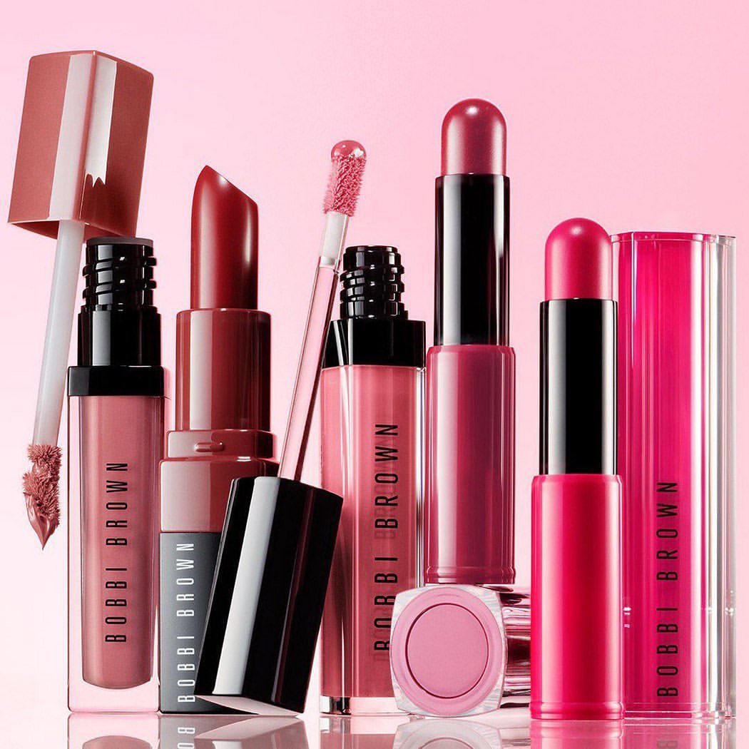 Cosmetics product still life: Assorted Bobbi Brown lipsticks and lip glosses in pink and red tones arranged on a light pink background.