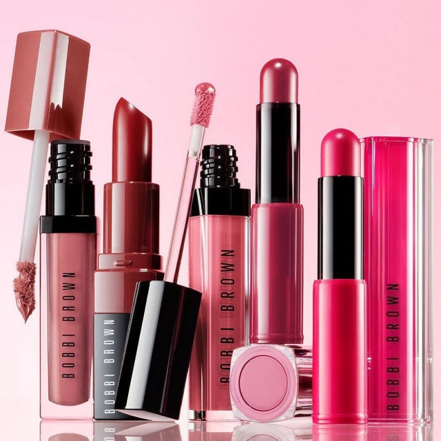 Cosmetics product still life: Assorted Bobbi Brown lipsticks and lip glosses in pink and red tones arranged on a light pink background.