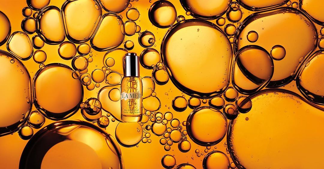 Close-up product shot of La Mer serum bottle amidst golden oil bubbles on a bright yellow background, showcasing skincare and luxury.