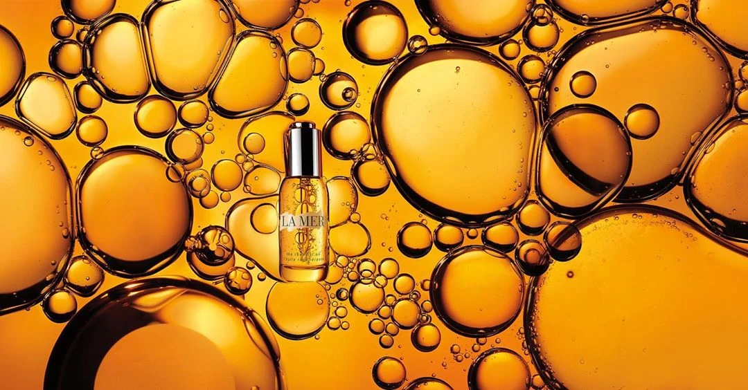Close-up product shot of La Mer serum bottle amidst golden oil bubbles on a bright yellow background, showcasing skincare and luxury.