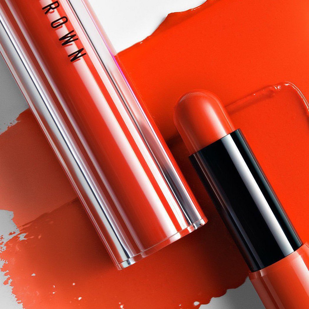 Close-up of Bobbi Brown lipstick in clear and black tube on a smear of orange lipstick against a white background, beauty product shot.