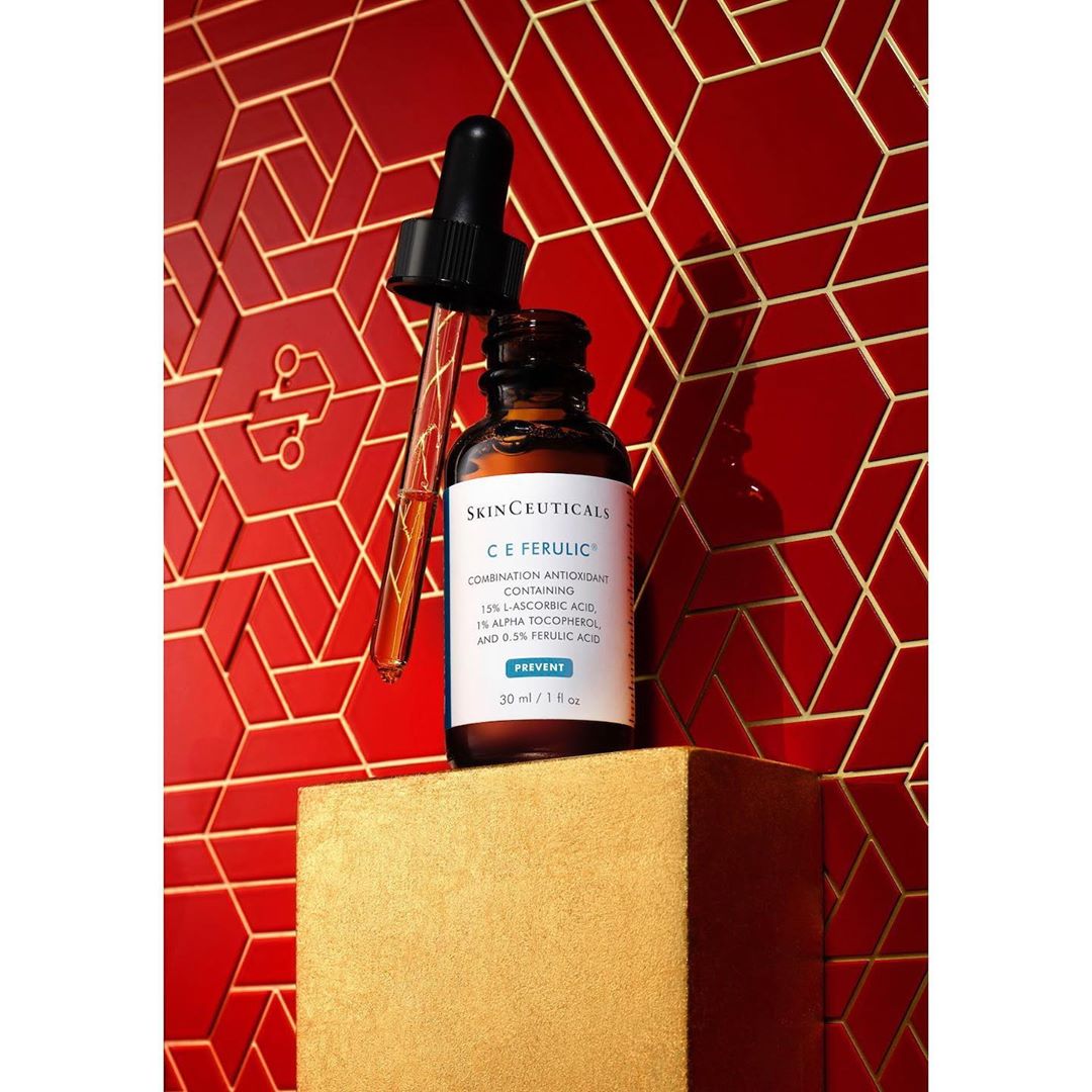 Still life of a skincare product, SkinCeuticals C E Ferulic serum, with an open dropper on a gold pedestal against a red geometric background.