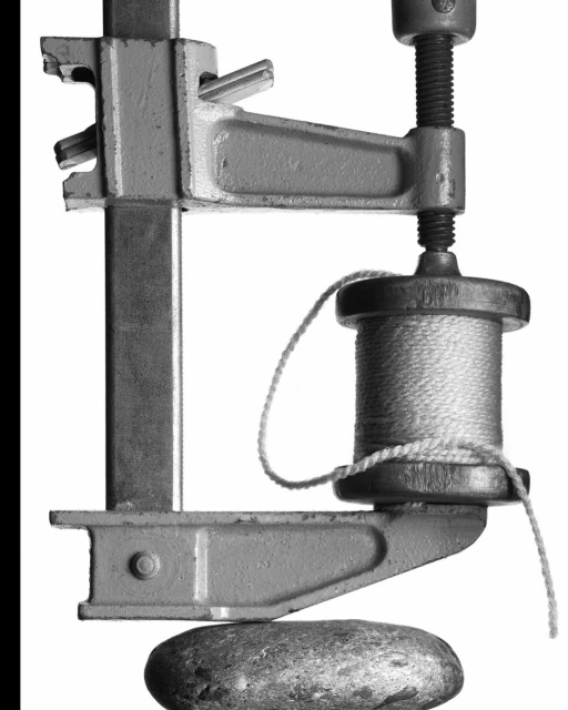 A monochrome still life features a metal clamp pressing a spool of thread against a smooth stone, highlighting textures and forms in stark light.