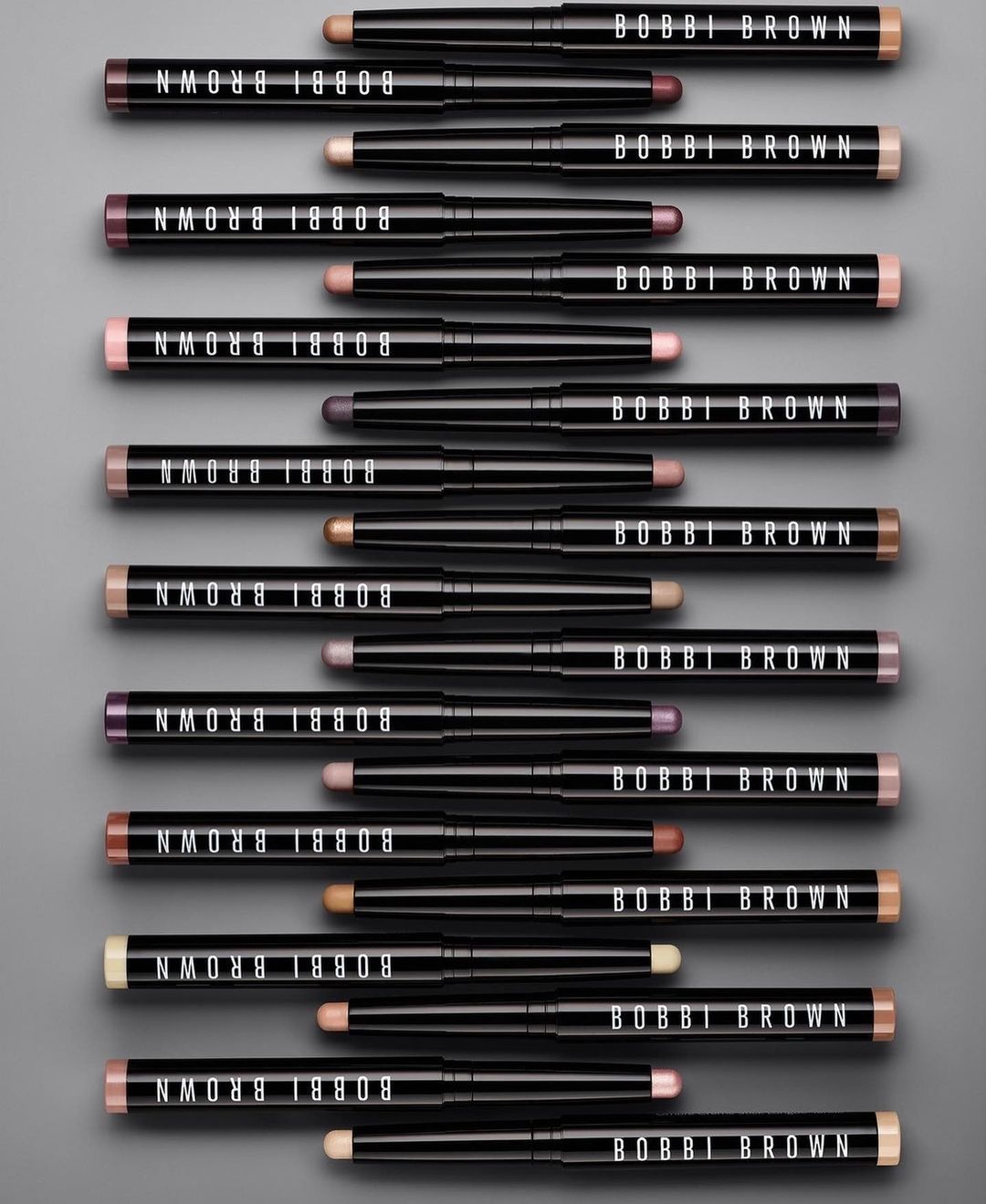 Bobbi Brown eyeshadow sticks in an array of colors arranged on a gray background. The black tubes feature the brand logo.