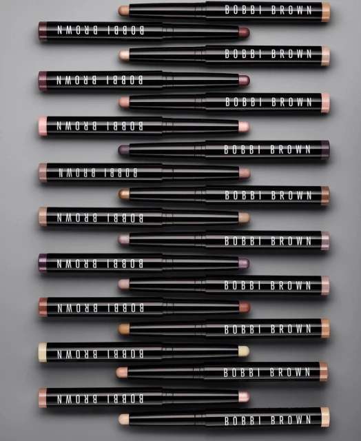 Bobbi Brown eyeshadow sticks in an array of colors arranged on a gray background. The black tubes feature the brand logo.