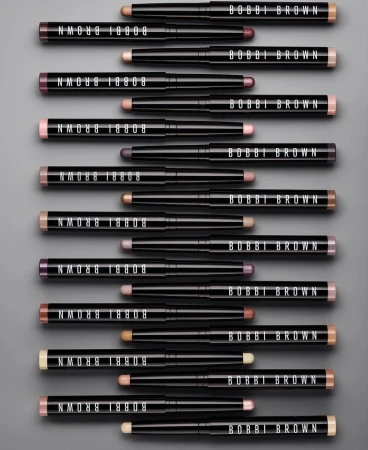 Bobbi Brown eyeshadow sticks in an array of colors arranged on a gray background. The black tubes feature the brand logo.