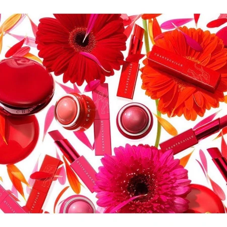 Chantecaille beauty products arranged with red and pink flowers, showcasing lipstick, blush, and powder compacts on white background.