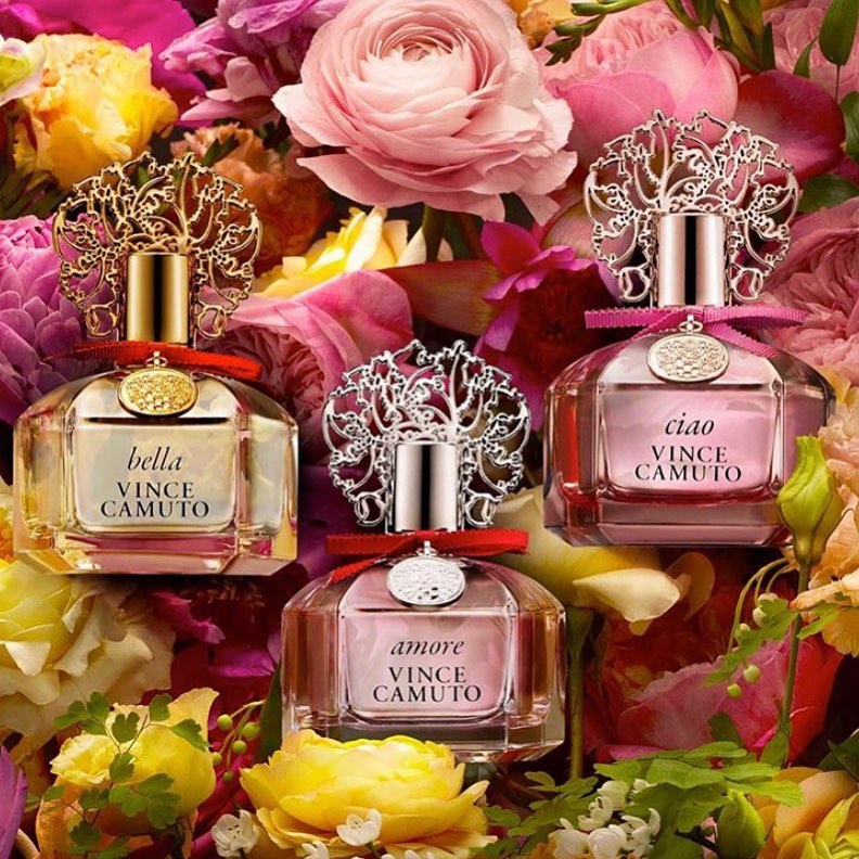 Three Vince Camuto perfume bottles displayed amidst a vibrant floral arrangement with pink, yellow, and purple blossoms.