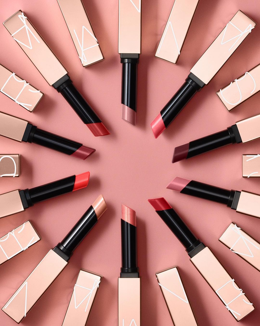 Overhead shot of NARS lipsticks arranged in a circle with alternating shades on a peach background, showcasing beauty products.