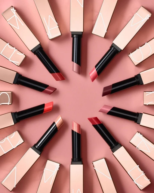 Overhead shot of NARS lipsticks arranged in a circle with alternating shades on a peach background, showcasing beauty products.