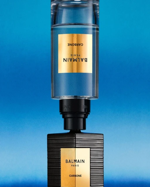 Two stacked Balmain Carbone perfume bottles are displayed against a vibrant blue background, emphasizing the brand's luxury and design.