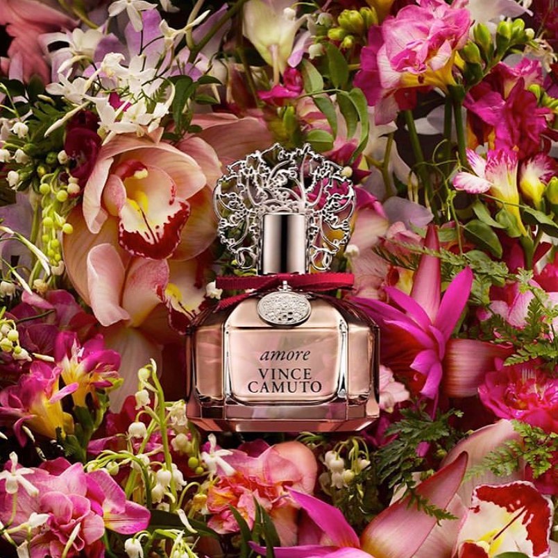 A bottle of Amore Vince Camuto perfume is set amidst a vibrant floral background, with pink, white, and purple blooms.