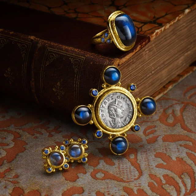 Close-up of gold earrings, ring, and pendant jewelry set featuring blue cabochon stones and set on an antique book with patterned backdrop.