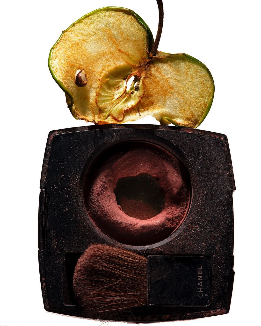 Still life of a half-eaten apple on top of a makeup compact with a brush, set against a stark white background.