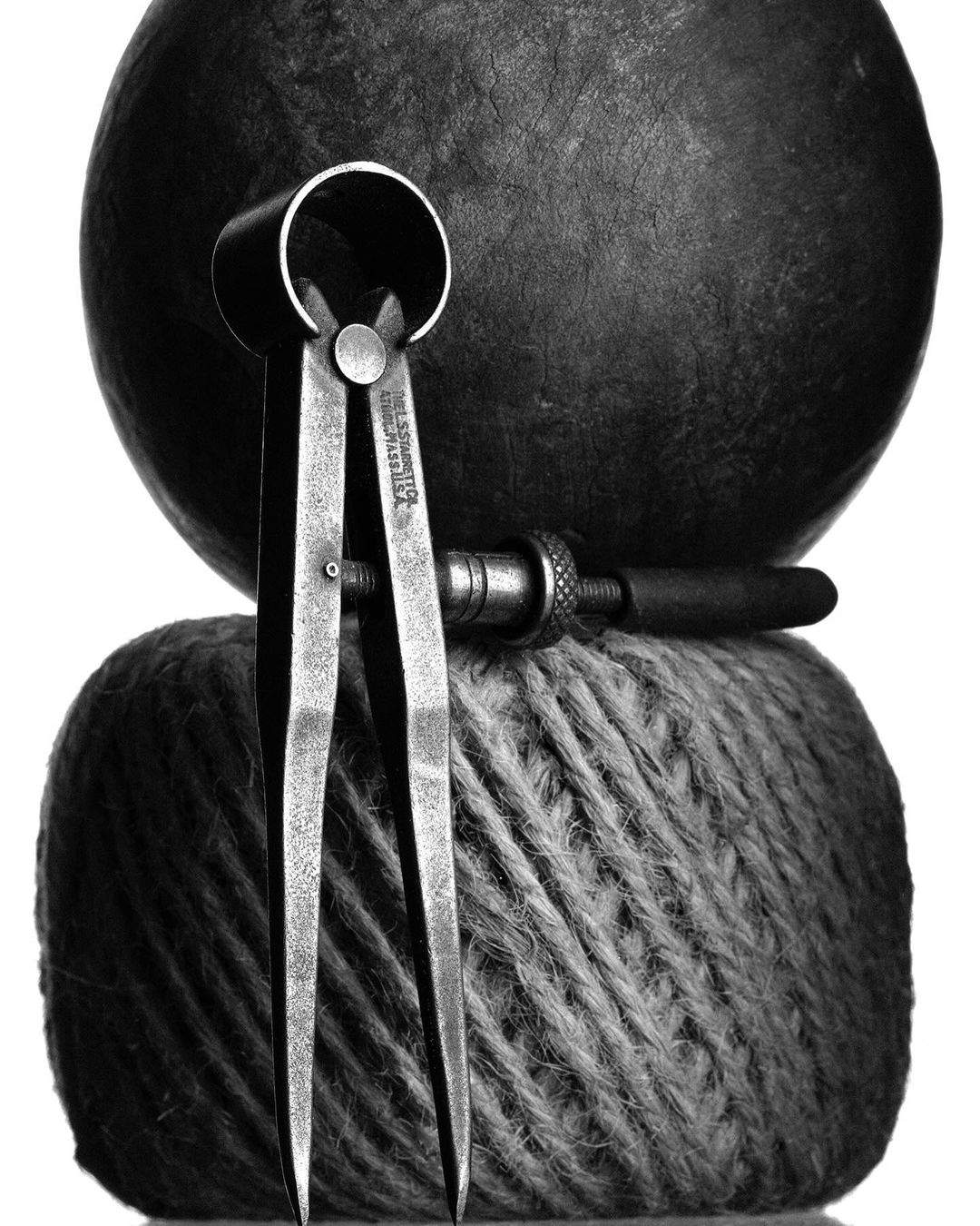Close-up black and white studio shot featuring metal calipers and a wood tool atop a textured ball of yarn, with a dark sphere in the background.