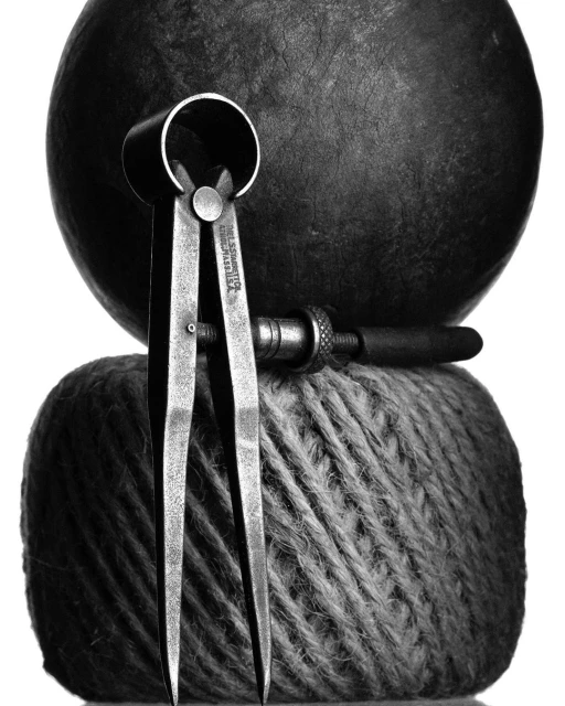 Close-up black and white studio shot featuring metal calipers and a wood tool atop a textured ball of yarn, with a dark sphere in the background.