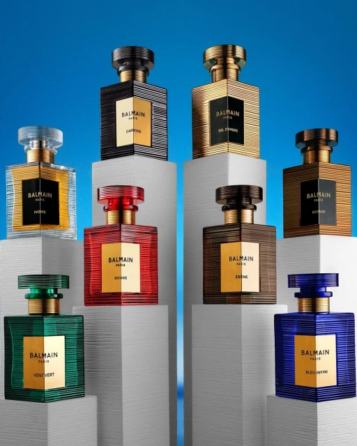 Collection of Balmain Paris perfume bottles in various colors on white pedestals against a gradient blue background. Fragrance product shot.
