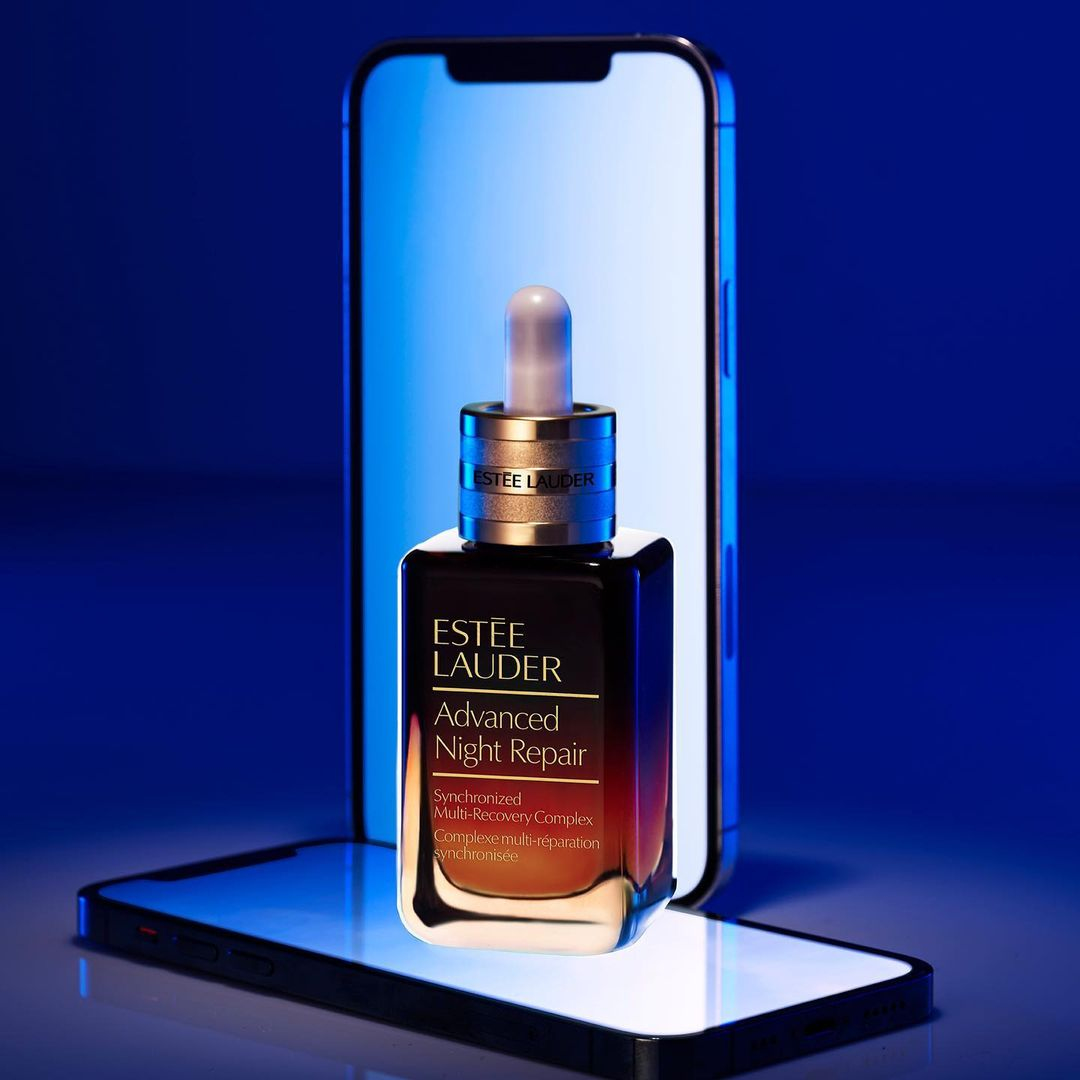 Estee Lauder Advanced Night Repair serum bottle stands on a smartphone in front of a larger illuminated phone screen in a photo studio.