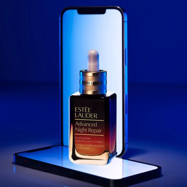 Estee Lauder Advanced Night Repair serum bottle stands on a smartphone in front of a larger illuminated phone screen in a photo studio.