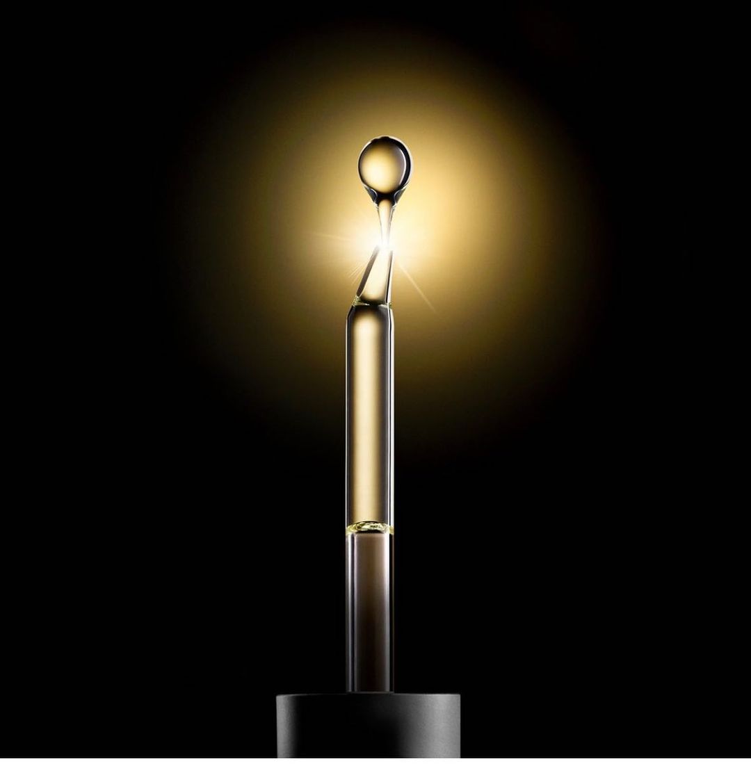 Gold cosmetic dropper bottle and bulb on a black background with halo lighting, offering a beauty or skincare product visual.