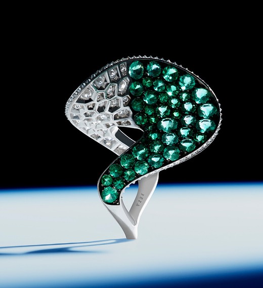 Close-up of a sculptural ring featuring green gemstones and white diamonds set in silver metal, presented against a dark background.