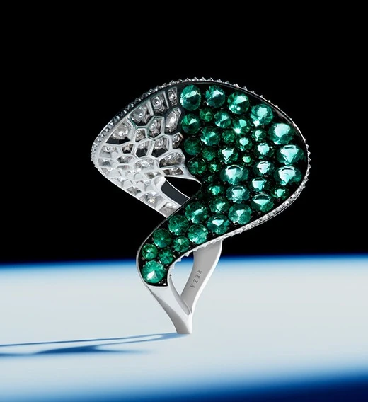 Close-up of a sculptural ring featuring green gemstones and white diamonds set in silver metal, presented against a dark background.