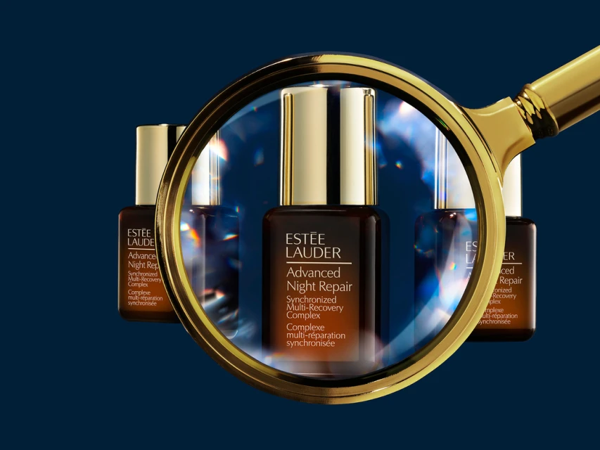 Three Estee Lauder Advanced Night Repair serum bottles, one magnified by a gold magnifying glass, on a gradient dark blue background.