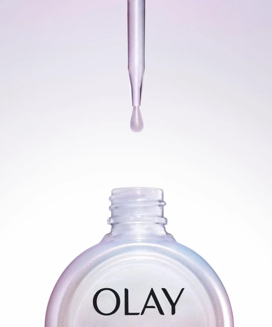 Close-up of a dropper dispensing liquid into a bottle of Olay skincare serum, set against a gradient white and purple background.