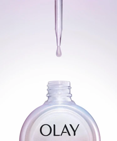 Close-up of a dropper dispensing liquid into a bottle of Olay skincare serum, set against a gradient white and purple background.