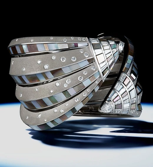 Close-up of an intricate, multi-tiered bracelet featuring silver metal, mother-of-pearl segments, and diamond accents against a dark backdrop.