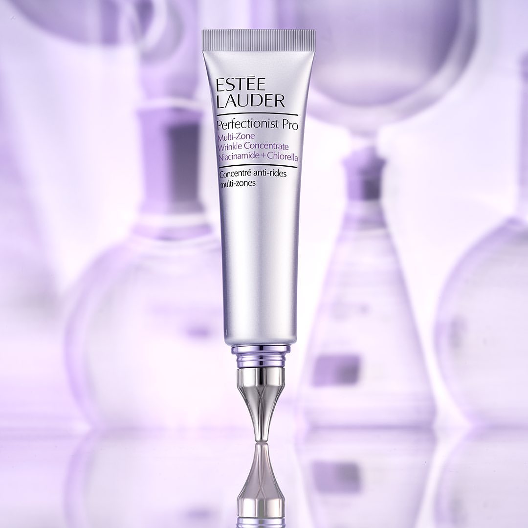 Estee Lauder Perfectionist Pro Multi-Zone Wrinkle Concentrate Niacinamide + Chlorella tube on a reflective surface with blurred flasks.