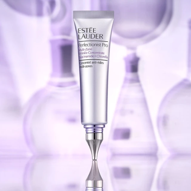 Estee Lauder Perfectionist Pro Multi-Zone Wrinkle Concentrate Niacinamide + Chlorella tube on a reflective surface with blurred flasks.